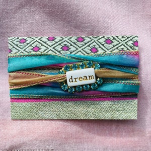 May include: A white card with a decorative design of pink and blue ribbons and a white button with the word "dream" in gold lettering.