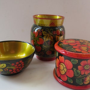 May include: Three hand-painted wooden nesting boxes with floral designs. The largest box is a vase with a gold rim and a floral pattern with red berries. The middle box is a bowl with a gold rim and a floral pattern with red berries. The smallest box is a round box with a red lid and a floral pattern with orange flowers.
