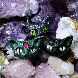 May include: Three cat head sculptures with glowing green eyes and a third eye, set among amethyst and other crystals, and animal skulls. The cat heads are black and green, with a mystical theme.