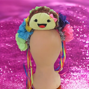 May include: A colorful flower crown with a smiling yellow plush face in the center. The crown is decorated with blue, pink, green, and yellow flowers, silver tinsel, and rainbow ribbons.