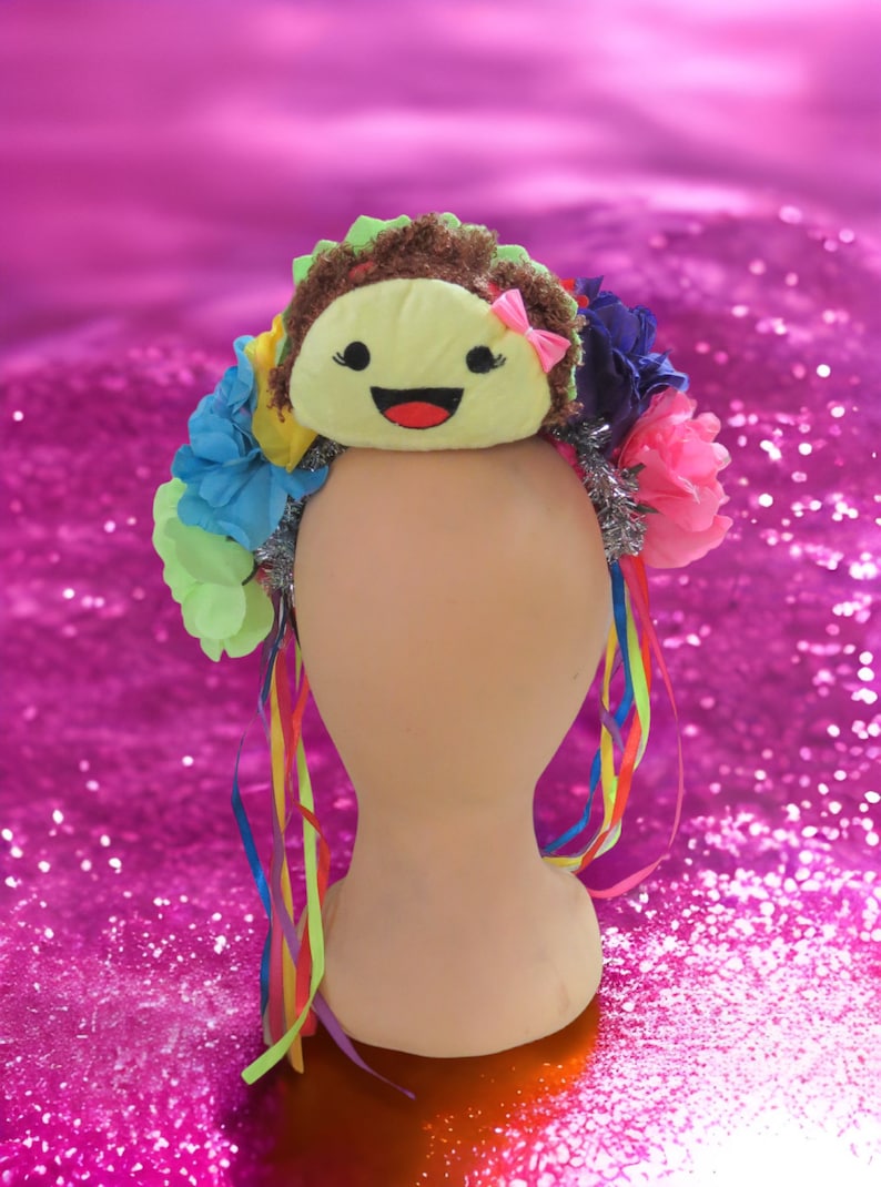 May include: A colorful flower crown with a smiling yellow plush face in the center. The crown is decorated with blue, pink, green, and yellow flowers, silver tinsel, and rainbow ribbons.