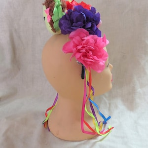 May include: A colorful flower crown headband with a green and brown plush bird on top. The headband has pink, purple, and red flowers with rainbow ribbons.