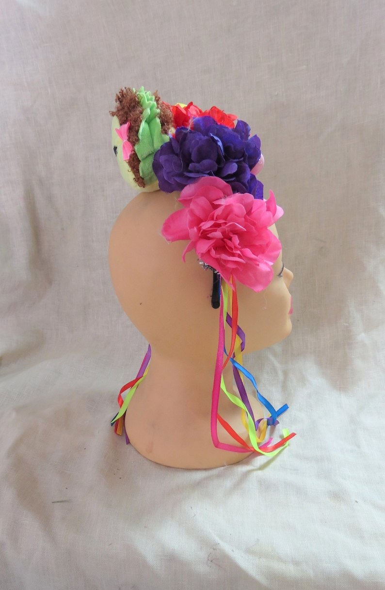 May include: A colorful flower crown headband with a green and brown plush bird on top. The headband has pink, purple, and red flowers with rainbow ribbons.