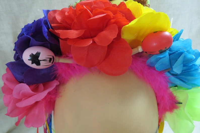 May include: A colorful flower crown with pink, red, yellow, blue, and purple flowers. The crown is decorated with maracas and feathers.