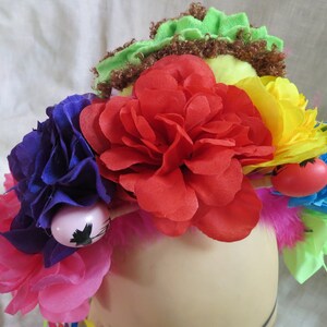 May include: A colorful flower crown with pink, red, yellow, blue, and green flowers. The crown is decorated with feathers and small red and pink balls.