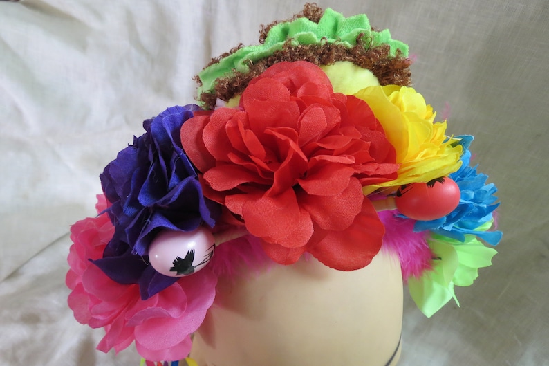 May include: A colorful flower crown with pink, red, yellow, blue, and green flowers. The crown is decorated with feathers and small red and pink balls.