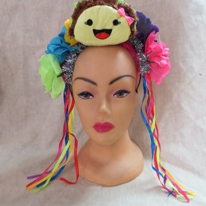 May include: A colorful headband with a yellow smiling face, pink flowers, green leaves, and silver tinsel. The headband has long rainbow ribbons attached.