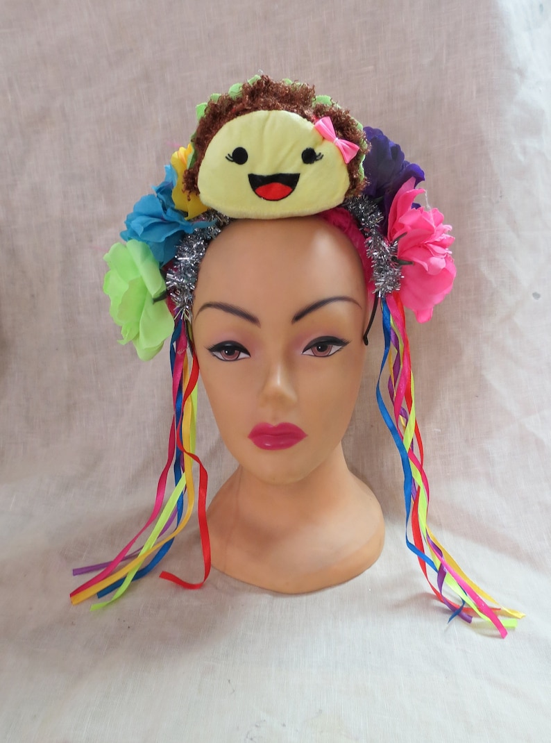 May include: A colorful headband with a yellow smiling face, pink flowers, green leaves, and silver tinsel. The headband has long rainbow ribbons attached.