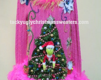 May include: Pink cable-knit Christmas sweater with a decorated tree and Grinch figure. The sweater has a pink feather trim and a pink and yellow feather collar. The website "tackyuglychristmassweaters.com" is printed on the sweater.