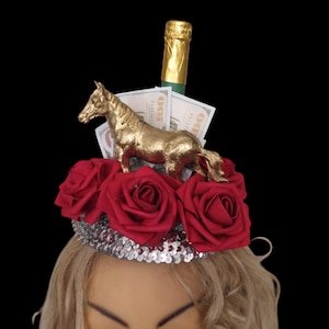 May include: A decorative headpiece featuring a gold horse figurine, red roses, and a bottle of champagne. The base is a silver sequined band. Hundred dollar bills are also included in the design.