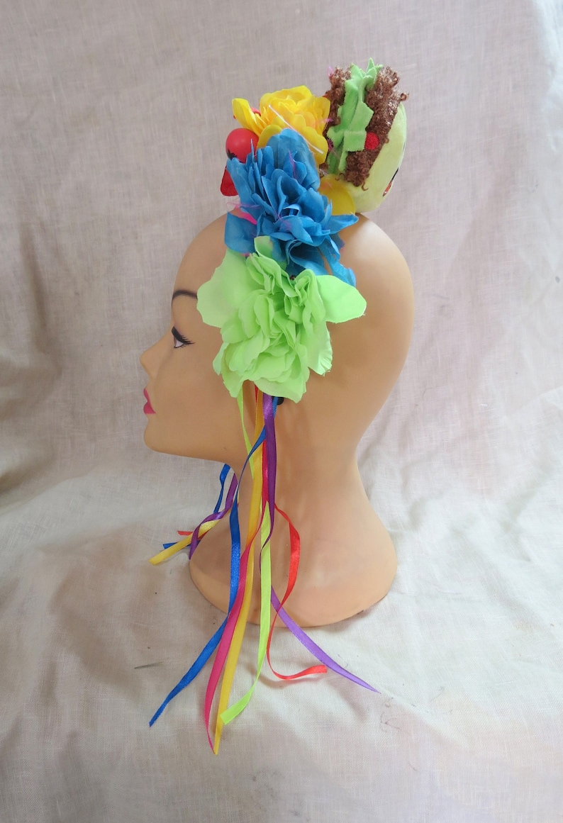 May include: A colorful flower crown with green, blue, yellow, and red flowers. The crown is decorated with a small stuffed green and brown figure. The crown is attached to a mannequin head with long, colorful ribbons.