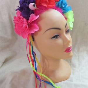 May include: A colorful flower crown with pink, red, purple, and green flowers. The crown has a pink base and is decorated with colorful ribbons.