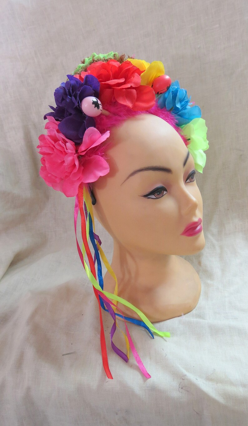 May include: A colorful flower crown with pink, red, purple, and green flowers. The crown has a pink base and is decorated with colorful ribbons.