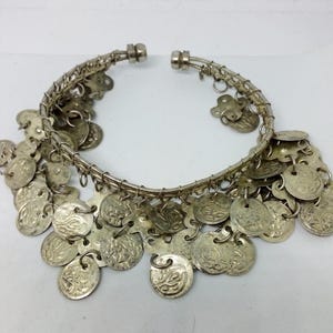 May include: Silver-toned bracelet with dangling coin charms. The bracelet features a wire-wrapped design and is open at the clasp. The coin charms have intricate embossed patterns. This jewelry piece is suitable for various occasions.