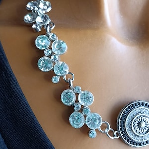 Può includere: Una collana color argento caratterizzata da una serie di cristalli trasparenti, rotondi e sfaccettati disposti a grappolo, collegata a un ciondolo circolare con un motivo floreale scuro, intricato e in rilievo e una catena a maglie.