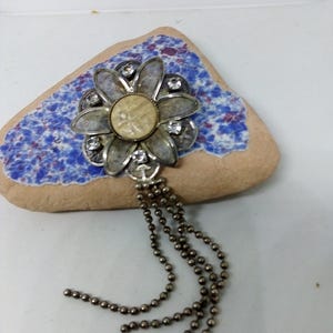 May include: A silver-toned flower brooch with a central beige disc and clear rhinestone accents. The brooch has layered petals and hangs from multiple strands of small, dark gray beads. The brooch is displayed on a small, blue and beige decorative stone.