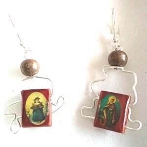 Op de afbeelding: Een paar handgemaakte oorbellen met religieuze afbeeldingen. Elke oorbel heeft een rode rechthoekige hanger met een ingelijste afbeelding van een heilige, een zilveren draadframe en een houten kraal. De oorbellen hangen aan zilveren haken.