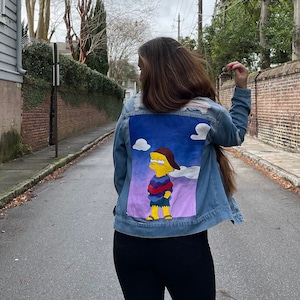 May include: A denim jacket with a painted image of a cartoon character on the back. The character is yellow with blue hair and is wearing a purple shirt and a blue hat. The background of the painting is a blue sky with white clouds.