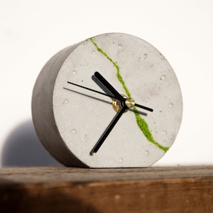 Peut inclure: Une horloge en béton gris avec des aiguilles noires et un motif de mousse verte. L'horloge est posée sur une surface en bois.