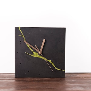 Peut inclure: Une horloge carrée noire avec des aiguilles dorées et un motif de mousse verte. L'horloge est posée sur une surface en bois.