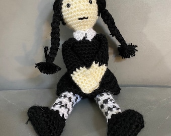 Könnte beinhalten: Eine gehäkelte Puppe mit schwarzen Haaren in Zöpfen, die ein schwarzes Kleid mit weißem Kragen und schwarz-weiß gestreiften Leggings trägt.