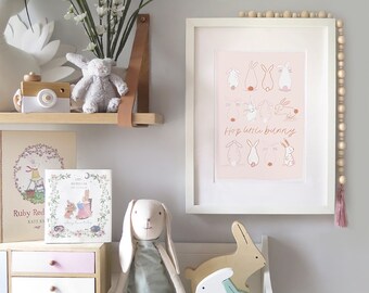 May include: A white framed print with a pink background featuring a series of illustrations of bunnies in different poses. The text "Hop little bunny" is written in brown cursive lettering. The print is displayed on a white shelf with a wooden bunny toy and a stuffed bunny toy.