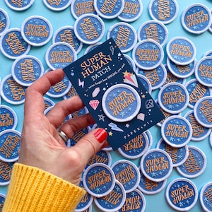 May include: A hand holds a card with the text "Super Human Patch" and the slogan "never forget how special you are". The card is surrounded by blue and white embroidered patches with the text "Super Human" on a light blue background.