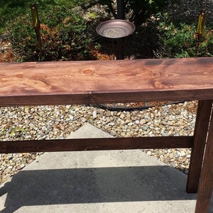 K&ouml;nnte beinhalten: Ein dunkelbrauner Holz-Konsolentisch mit schlichtem Design. Der Tisch hat eine rechteckige Tischplatte und zwei Beine auf jeder Seite. Der Tisch ist aus massivem Holz gefertigt und hat eine nat&uuml;rliche Oberfl&auml;che.
