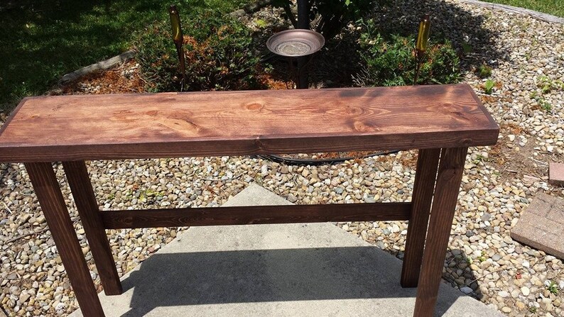 K&ouml;nnte beinhalten: Ein dunkelbrauner Holz-Konsolentisch mit schlichtem Design. Der Tisch hat eine rechteckige Tischplatte und zwei Beine auf jeder Seite. Der Tisch ist aus massivem Holz gefertigt und hat eine nat&uuml;rliche Oberfl&auml;che.