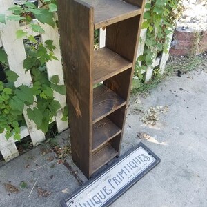 May include: A dark brown wooden shelf unit with four shelves. The shelf unit is standing on a gray concrete surface. A black and white sign with the text "Rustic Farmhouse Furniture Unique Primitives" is lying on the ground in front of the shelf unit.