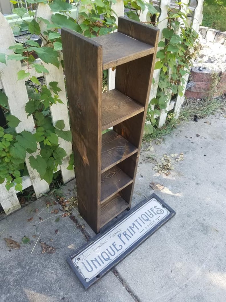 May include: A dark brown wooden shelf unit with four shelves. The shelf unit is standing on a gray concrete surface. A black and white sign with the text "Rustic Farmhouse Furniture Unique Primitives" is lying on the ground in front of the shelf unit.