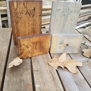 May include: Two wooden phone stands, one stained brown and one painted gray, are sitting on a wooden surface.  The stands are both rectangular and have a small shelf on the bottom.