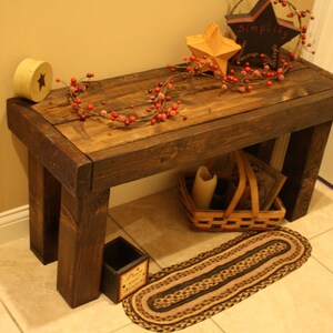 May include: A rustic, dark-stained wooden bench with a slatted top. The bench features a star-shaped decoration, a small basket, and a decorative garland. A small rug is placed in front of the bench.