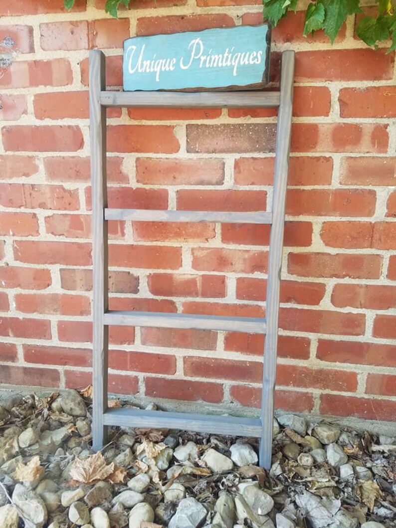 May include: A gray wooden ladder shelf with four shelves. The shelf has a sign that says "Unique Primtiques" in white lettering on a light blue background.