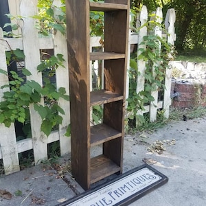 May include: A dark brown wooden shelf unit with four shelves. The shelf unit is standing on a gray concrete surface. A black sign with white text reads "Rustic Farmhouse Furniture Unique Primitives".