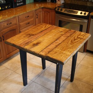 Puede incluir: Una mesa de madera r&uacute;stica con un acabado de madera natural y patas pintadas de negro. La mesa es cuadrada y tiene un dise&ntilde;o sencillo. Est&aacute; sentada en un suelo de baldosas.