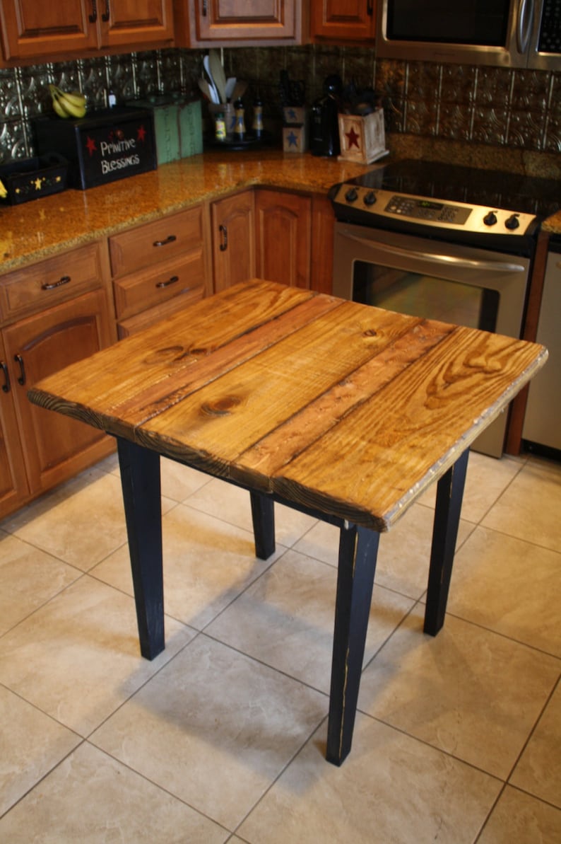 Puede incluir: Una mesa de madera r&uacute;stica con un acabado de madera natural y patas pintadas de negro. La mesa es cuadrada y tiene un dise&ntilde;o sencillo. Est&aacute; sentada en un suelo de baldosas.