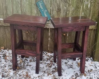 May include: Two dark brown wooden stools with a simple design. The stools have a rectangular seat and a single shelf underneath. The stools are sitting on a snowy ground with brown leaves.