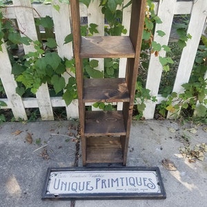 May include: A rustic wooden shelf unit with four shelves. The shelf unit is standing on a concrete surface. A sign with the text "Rustic Farmhouse Furniture Unique Primitives & Decor" is lying on the ground in front of the shelf unit.