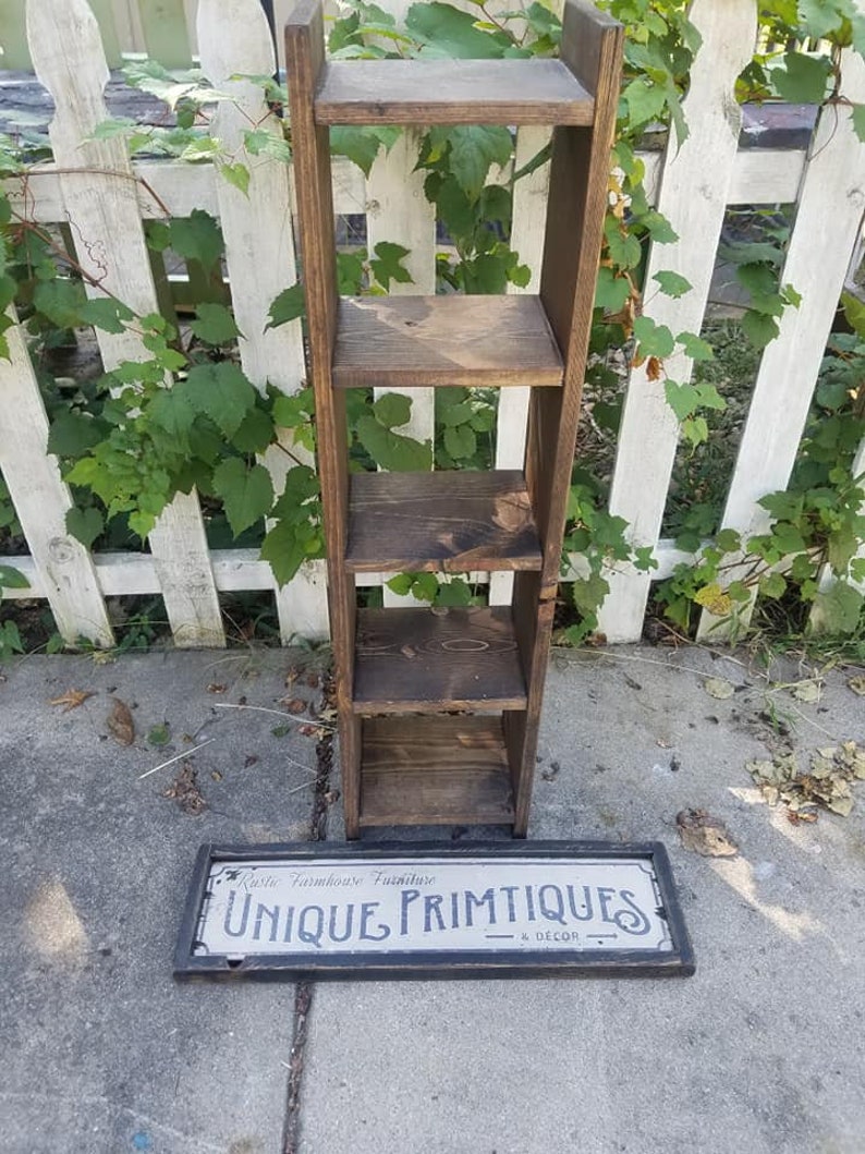 May include: A rustic wooden shelf unit with four shelves. The shelf unit is standing on a concrete surface. A sign with the text "Rustic Farmhouse Furniture Unique Primitives & Decor" is lying on the ground in front of the shelf unit.