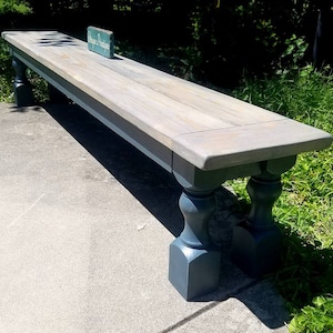 May include: A long wooden bench with a gray wash finish and dark gray turned legs. The bench has a sign on top that reads "Unique Designs".