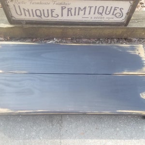 May include: A black wooden step stool with distressed paint and a rustic farmhouse style. The stool is made of two pieces of wood that are joined together. The top of the stool is slightly wider than the bottom. The stool is sitting on a gray concrete surface.