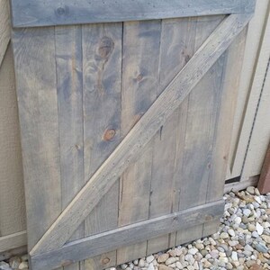 May include: A gray wooden door with a diagonal brace and a horizontal brace across the top. The door is made of reclaimed wood and has a rustic look.