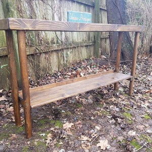 May include: A rustic wooden console table with a single shelf. The table is stained a dark brown color and has a simple, rectangular design. The table is sitting on a bed of brown leaves and moss.