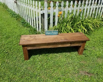 May include: A rectangular wooden bench with a dark brown stained finish and a smooth flat seat. Resting on the center of the bench is a small rectangular teal sign with the white text Unique Primtiques printed in a cursive script font.