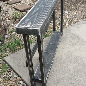 May include: A black wooden console table with a distressed finish and a lower shelf. The table is standing on a gravel surface.