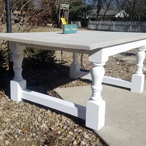 May include: A large square dining table featuring a light gray wooden tabletop and a white base with four thick, turned legs connected by horizontal support beams. A small teal sign with the white text Unique Prantique sits on the table surface.