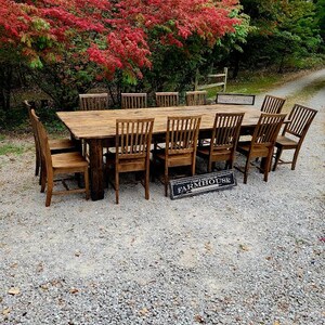 May include: A large wooden farmhouse table with a rustic finish, surrounded by chairs. The table is set in a gravel driveway with a backdrop of fall foliage.