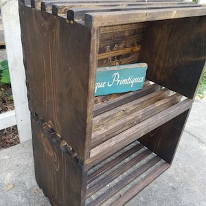 May include: A dark brown wooden shelf unit with three shelves. The shelves are made of slats of wood and the unit is stacked on top of itself. A small blue sign with the text "que Primitiques" is on the top shelf.
