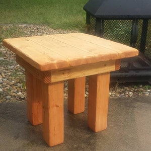 May include: A small, wooden end table with a natural wood finish. The table has a square top and four legs. The table is sitting on a concrete surface.